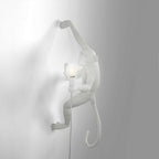 This white Clérachic monkey light climbs a grey-white wall, one arm stretched up, with a softly glowing bulb in its hand.