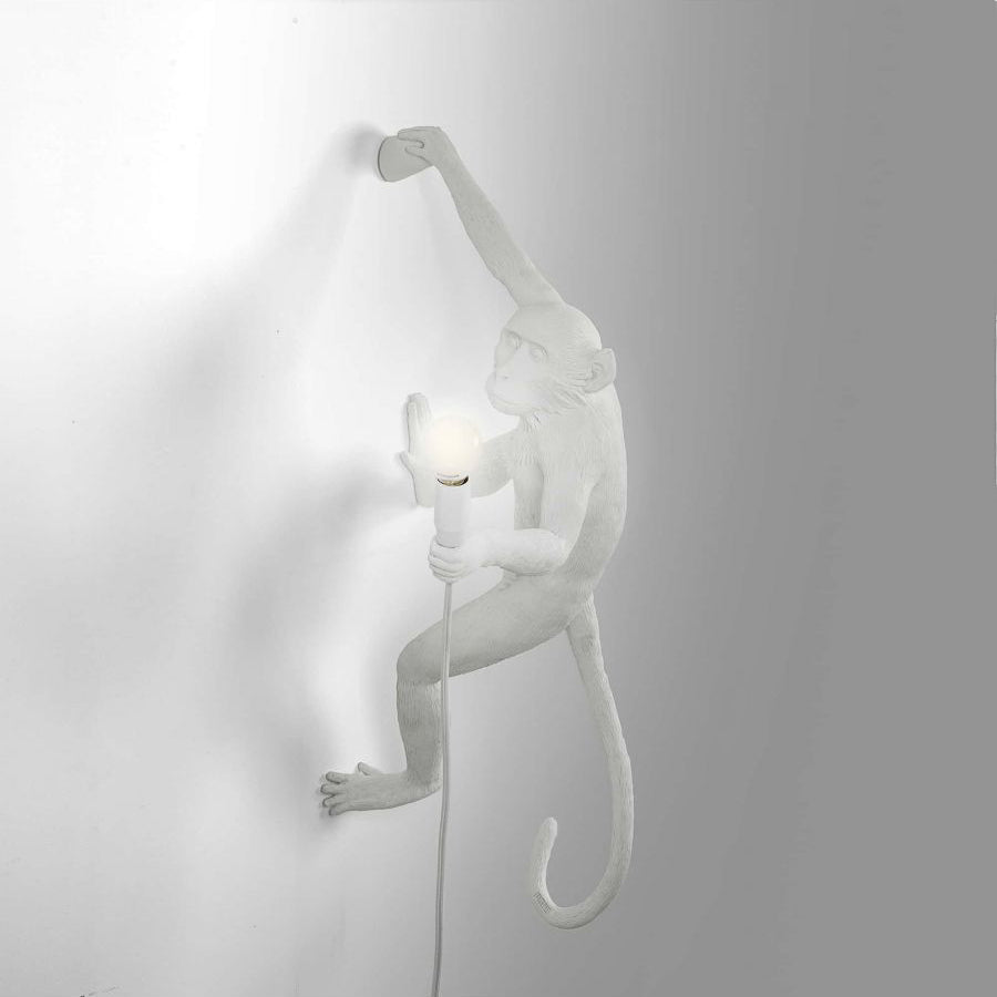 This white Clérachic monkey light climbs a grey-white wall, one arm stretched up, with a softly glowing bulb in its hand.