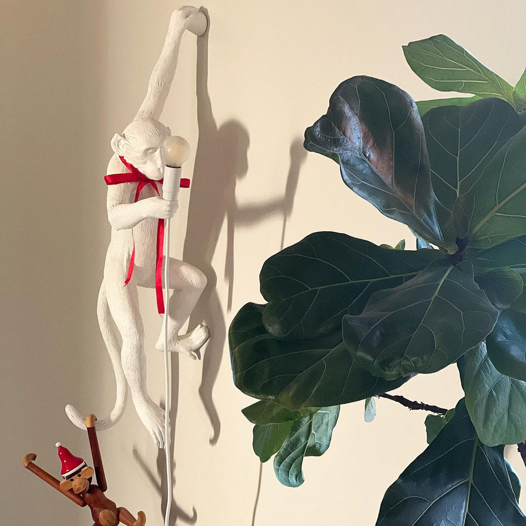 A white Clérachic monkey light with a red ribbon round its neck hangs beside a large fiddle-leaf fig on a cream wall.