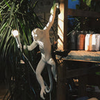 White monkey wall light from Clérachic reaching out among tall palms, with a wooden shelf of jars and a lit bulb in hand.
