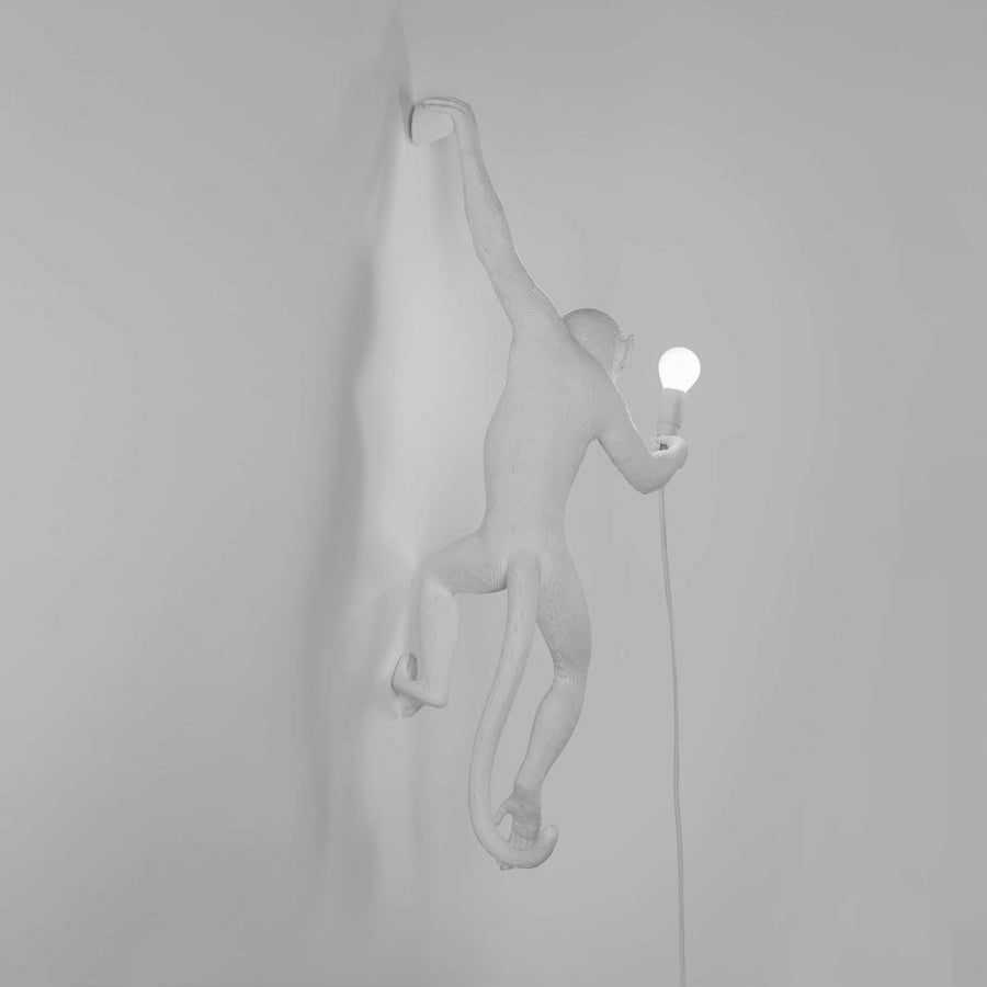 Soft grey view of a white Clérachic monkey light on a matching wall, one arm gripping the top and a small bulb lit.