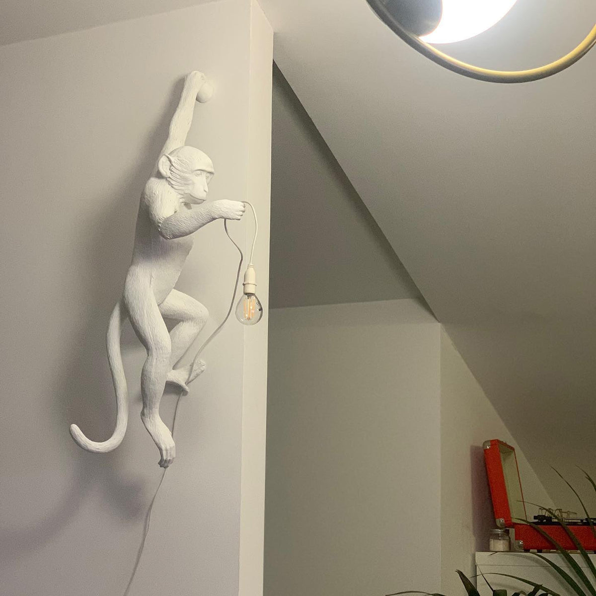 Beside a sloping ceiling and a pendant ring, a white Clérachic monkey wall light hangs with its bulb dangling on a short cord.