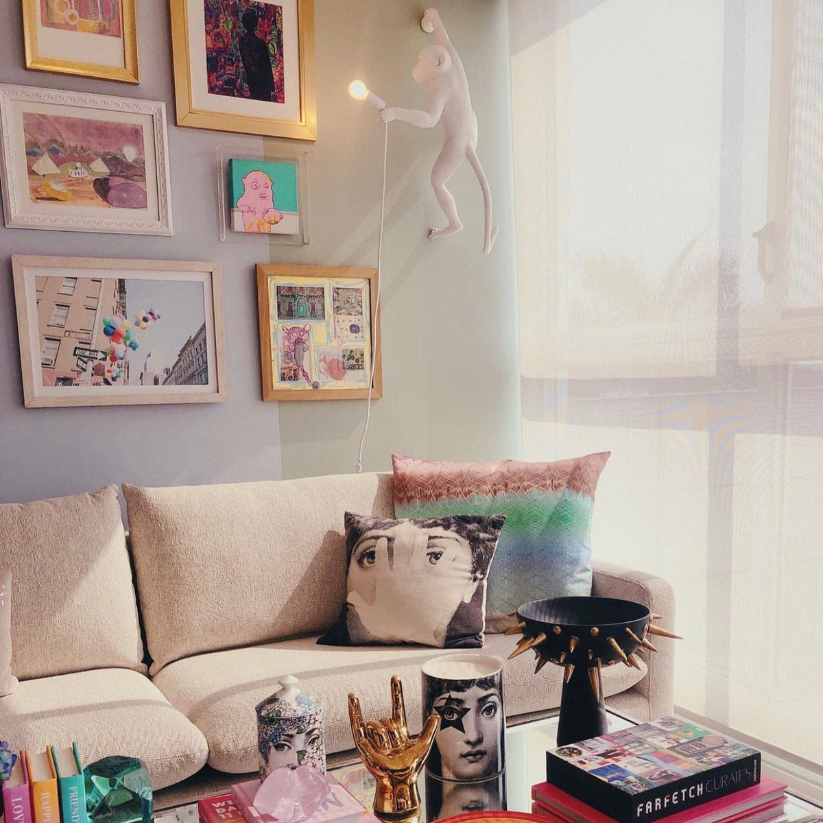 A white Clérachic monkey light sits high on a lilac-grey wall above a cream sofa, among colourful framed pictures.
