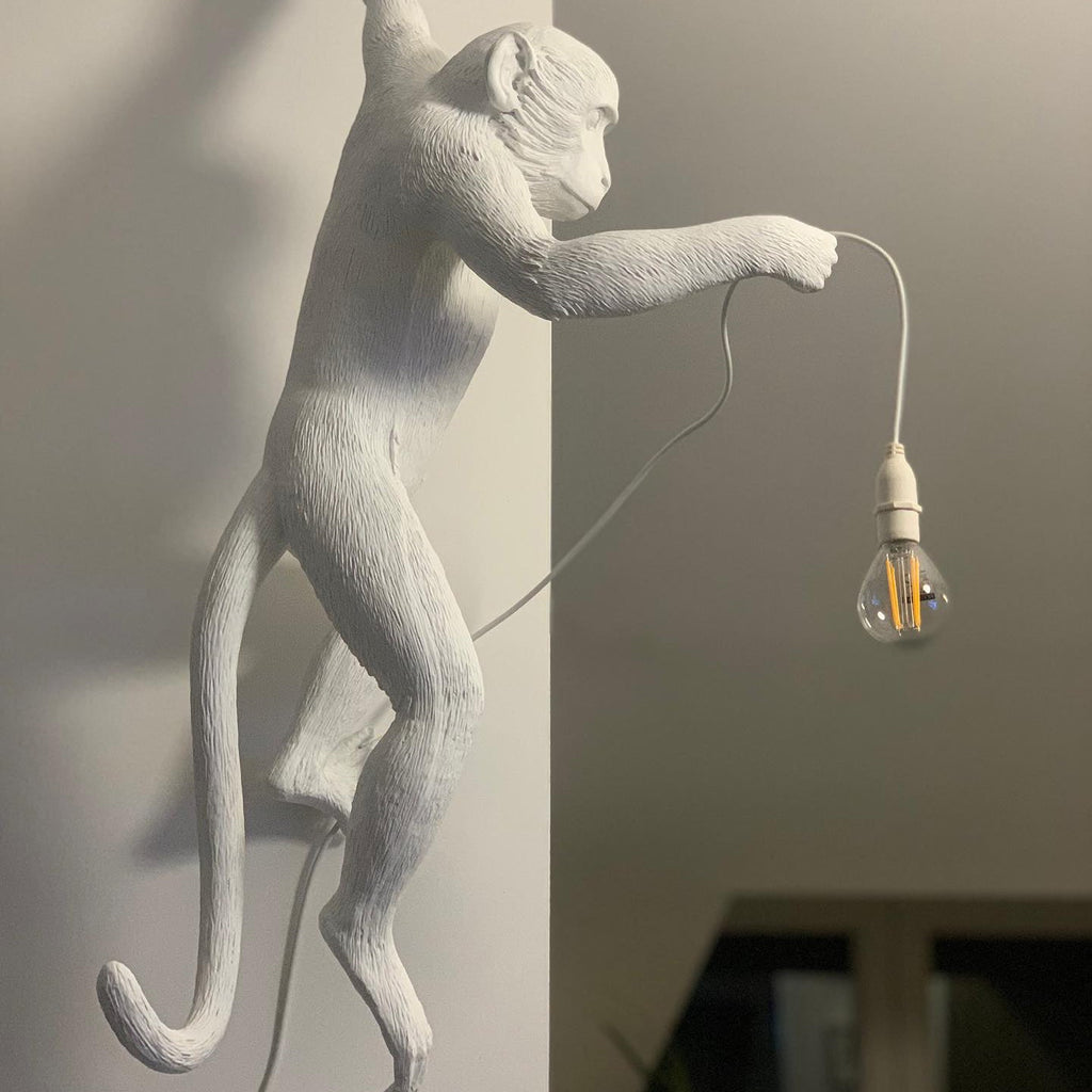 Close view of the white resin monkey on this Clérachic wall light, one arm out holding a filament bulb on a white cord.