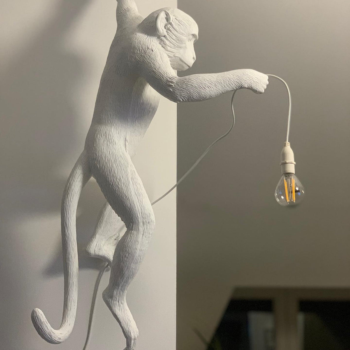Close view of the white resin monkey on this Clérachic wall light, one arm out holding a filament bulb on a white cord.