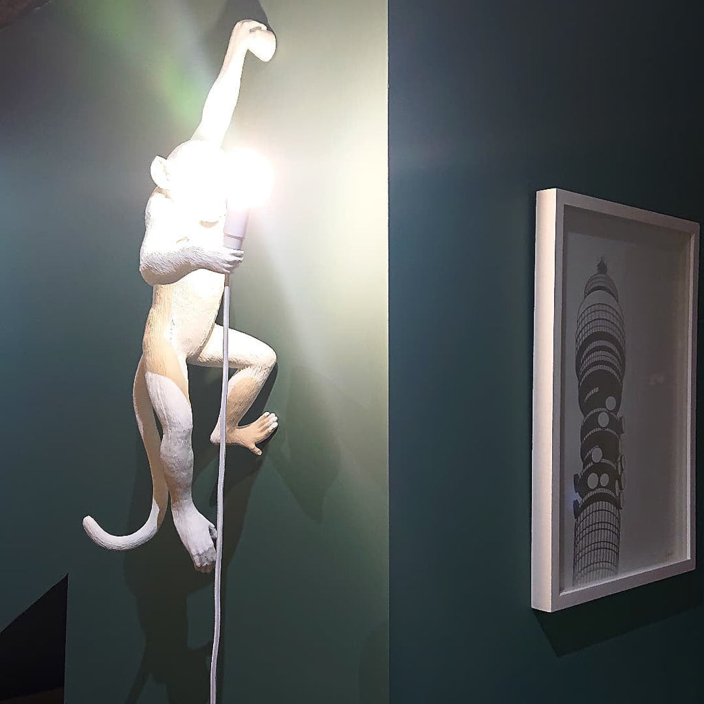 A white Clérachic monkey light, its bulb glowing brightly, climbs a dark teal wall beside a white framed print.