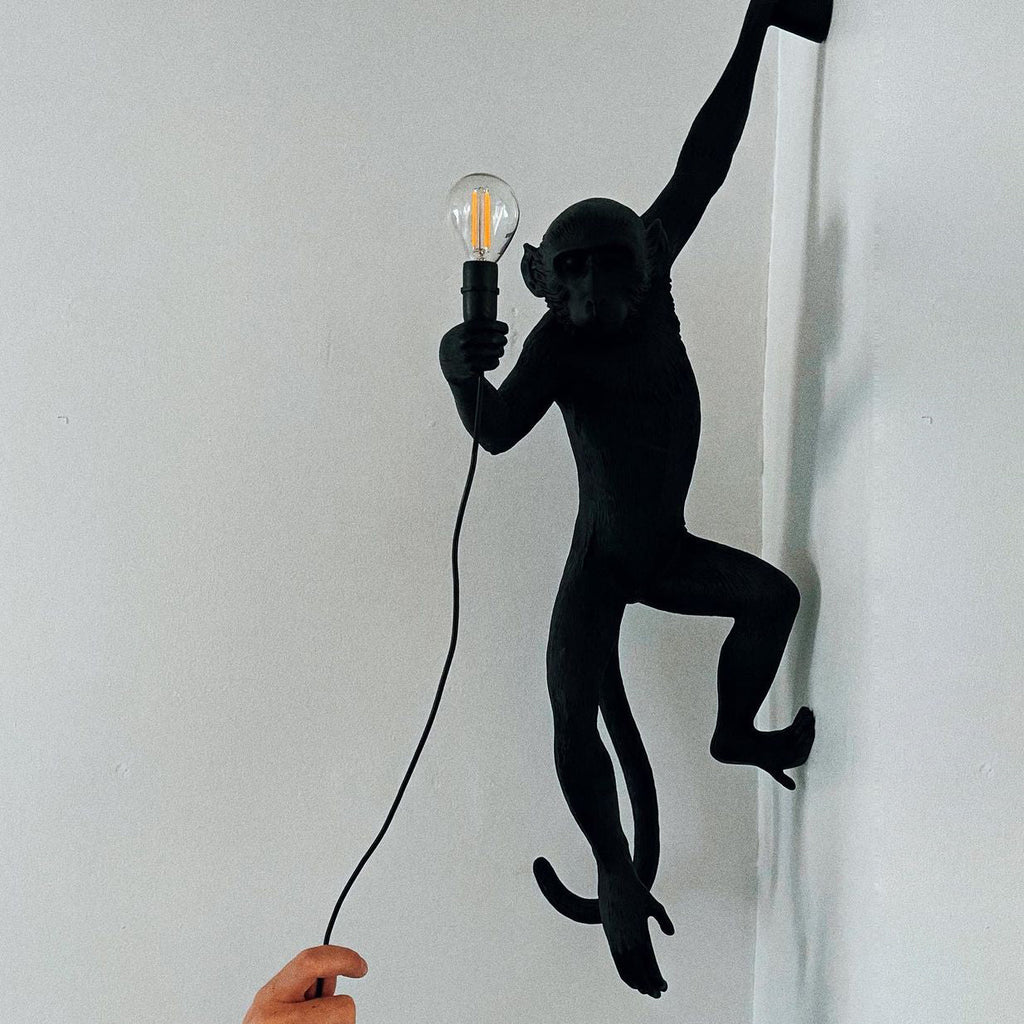 Black monkey wall light from Clérachic, close up on a pale wall, holding a filament bulb with its cord falling to a hand below.