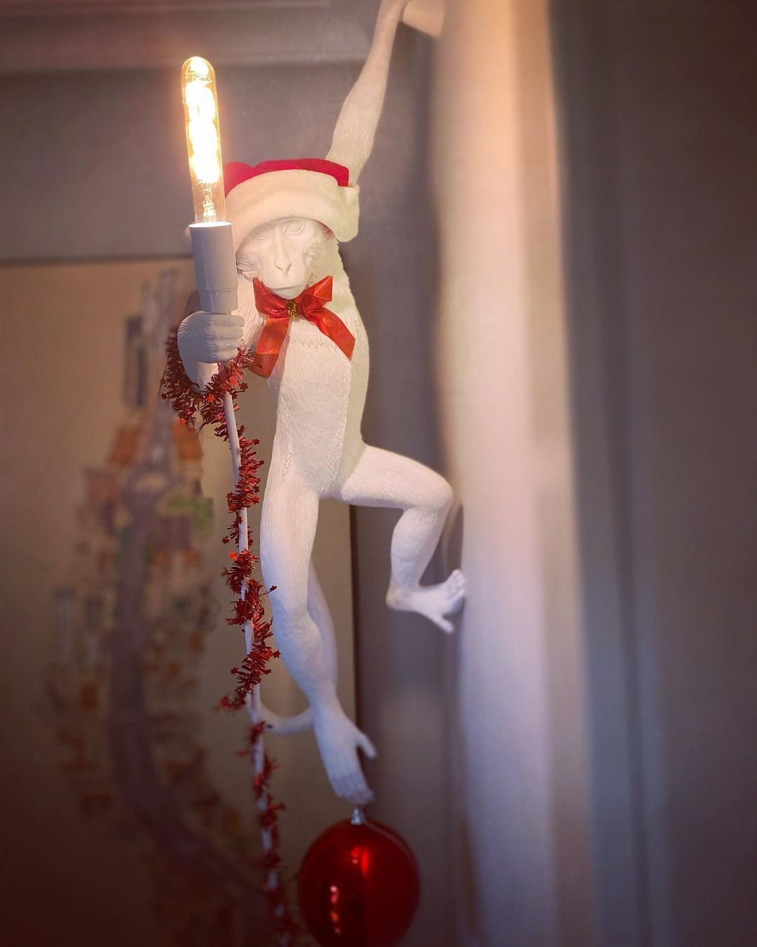 A white Clérachic monkey light wearing a red Santa hat and bow, with red tinsel wound around its arm and a tall bulb lit.