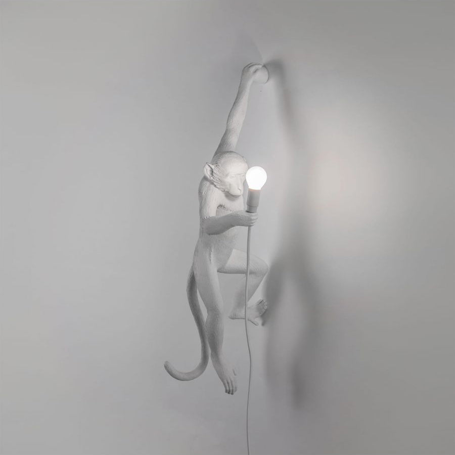 A white Clérachic monkey wall light fades into a pale grey wall, one arm raised, with a round bulb lit at the right.