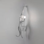 A white Clérachic monkey wall light fades into a pale grey wall, one arm raised, with a round bulb lit at the right.