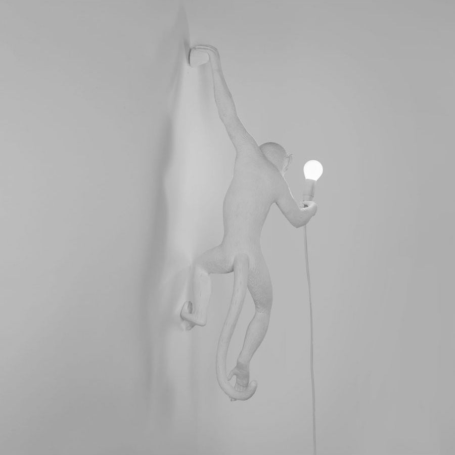 White resin monkey of this Clérachic wall light, shown in side light on a grey wall with a small lit bulb in its raised hand.