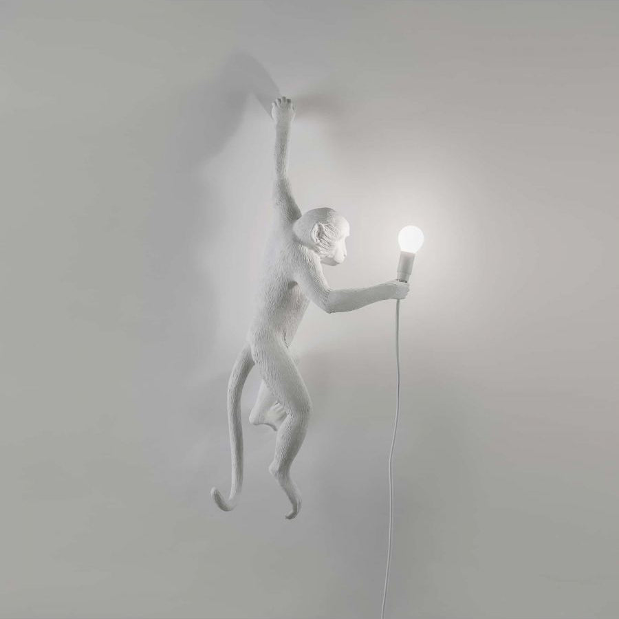A white Clérachic monkey light climbs a plain grey wall, its shadow soft beneath, holding a round bulb on a thin white cord.