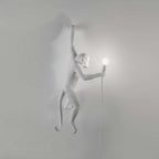 A white Clérachic monkey light climbs a plain grey wall, its shadow soft beneath, holding a round bulb on a thin white cord.