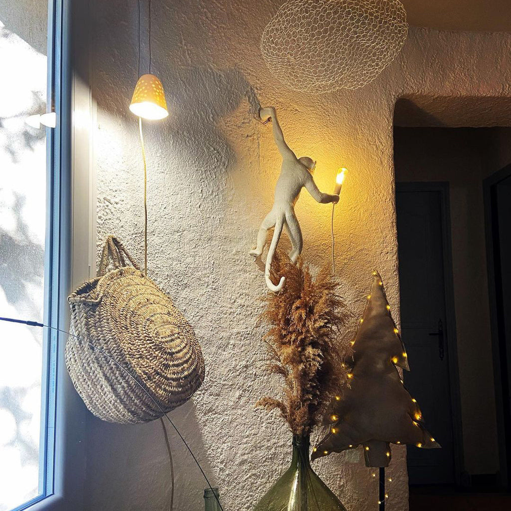 A white Clérachic monkey wall light glows warmly on a rough plaster wall beside a woven basket, pampas grass and a window.