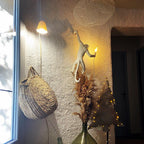 A white Clérachic monkey wall light glows warmly on a rough plaster wall beside a woven basket, pampas grass and a window.