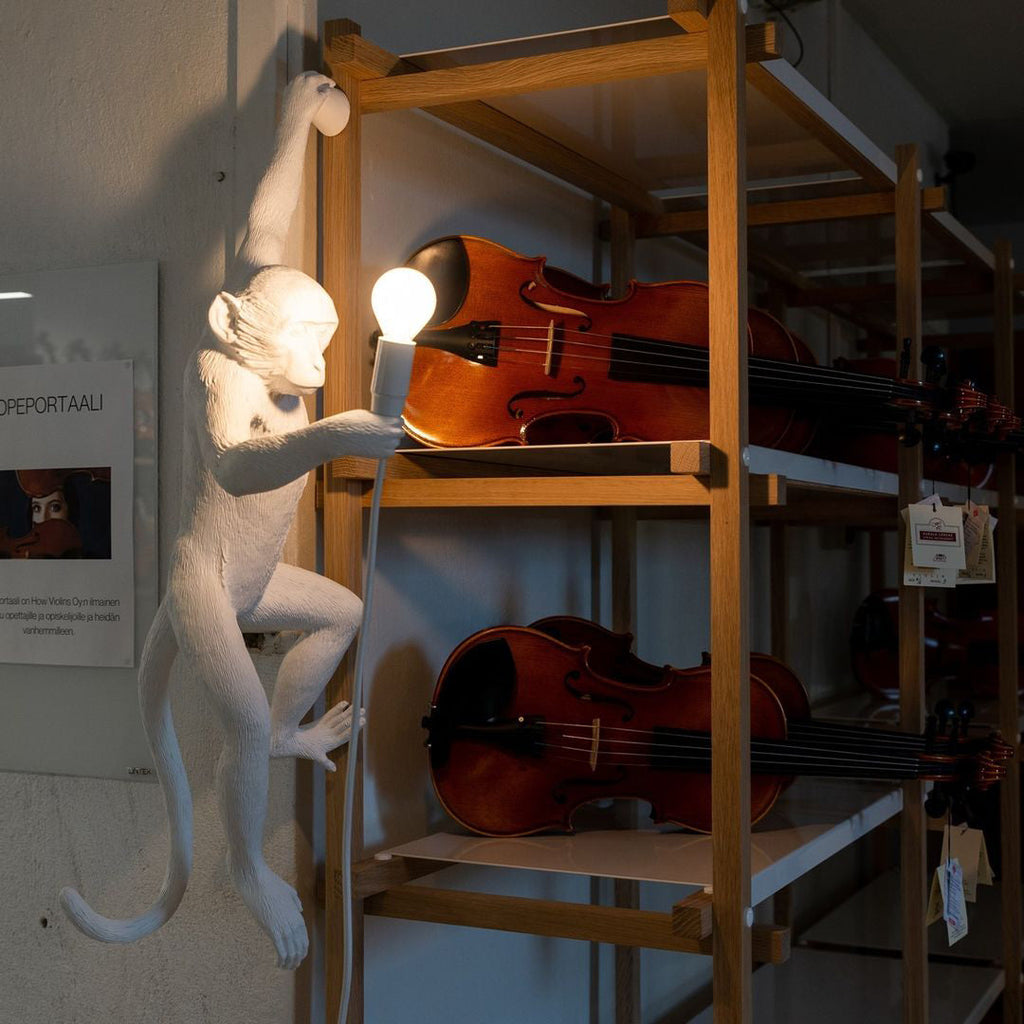 White monkey wall light from Clérachic holding a bulb beside wooden shelves, with violins on the shelf behind it.