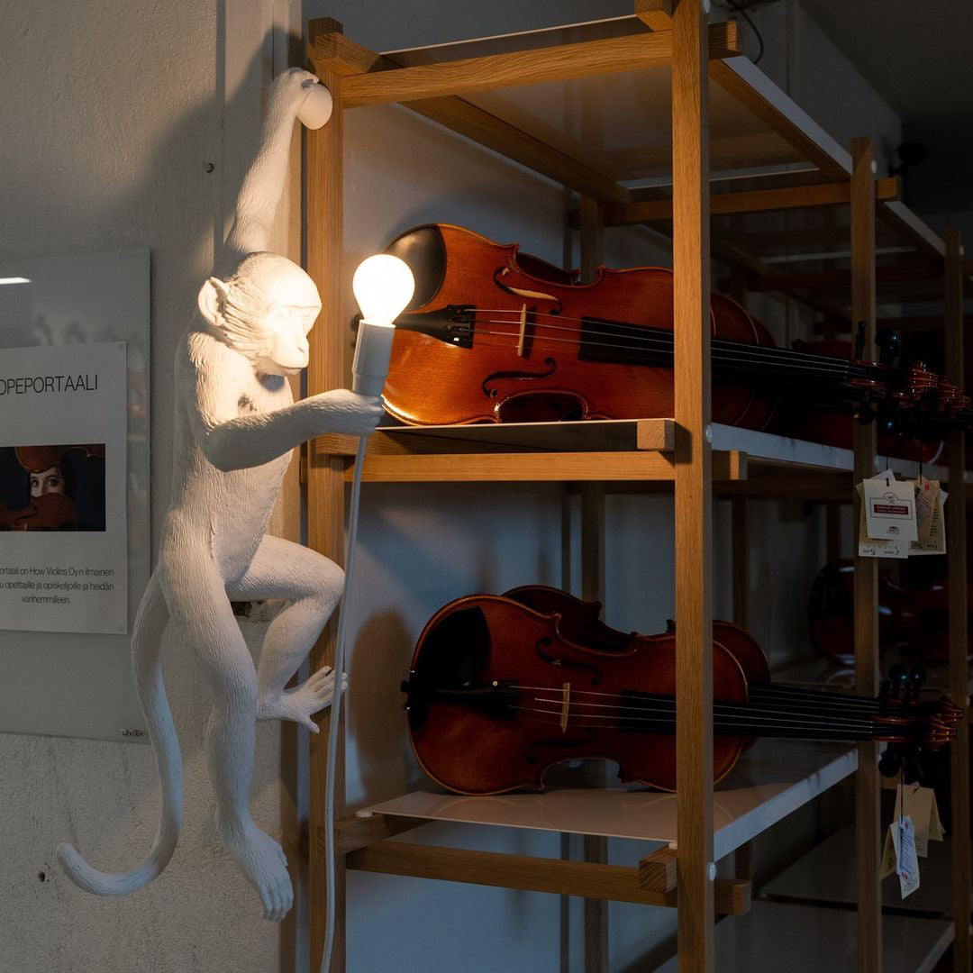 White monkey wall light from Clérachic holding a bulb beside wooden shelves, with violins on the shelf behind it.