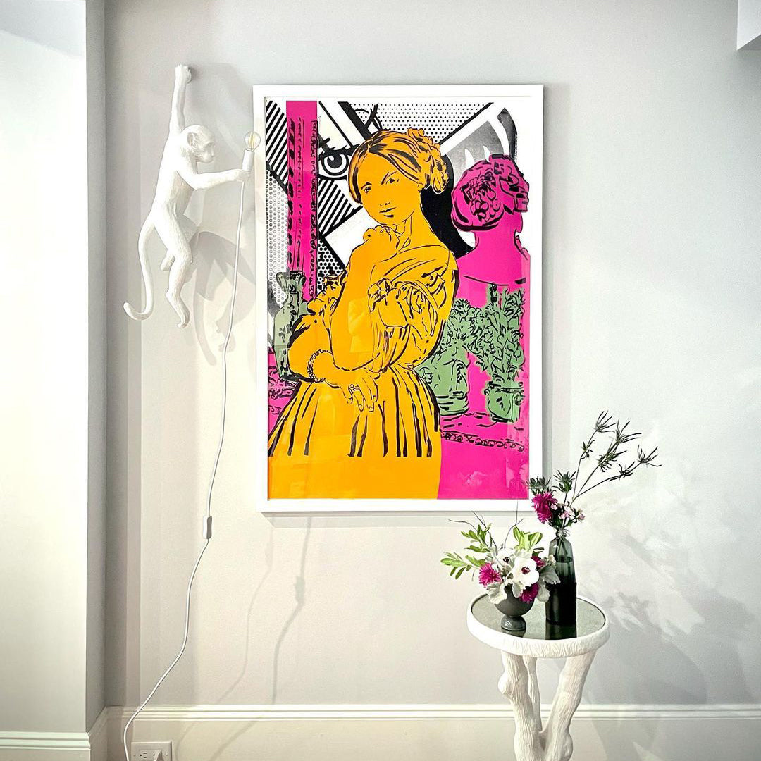 Far above a console table with a vase of flowers, a white Clérachic monkey wall light hangs beside a bold pink-and-yellow print.