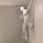 Close view of a white Clérachic monkey light on a grey wall, one arm reaching up, a round bulb held near its face.