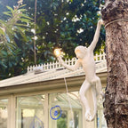 A white Clérachic monkey wall light hangs from a tree trunk in a garden, a lit bulb in hand and a glasshouse behind.