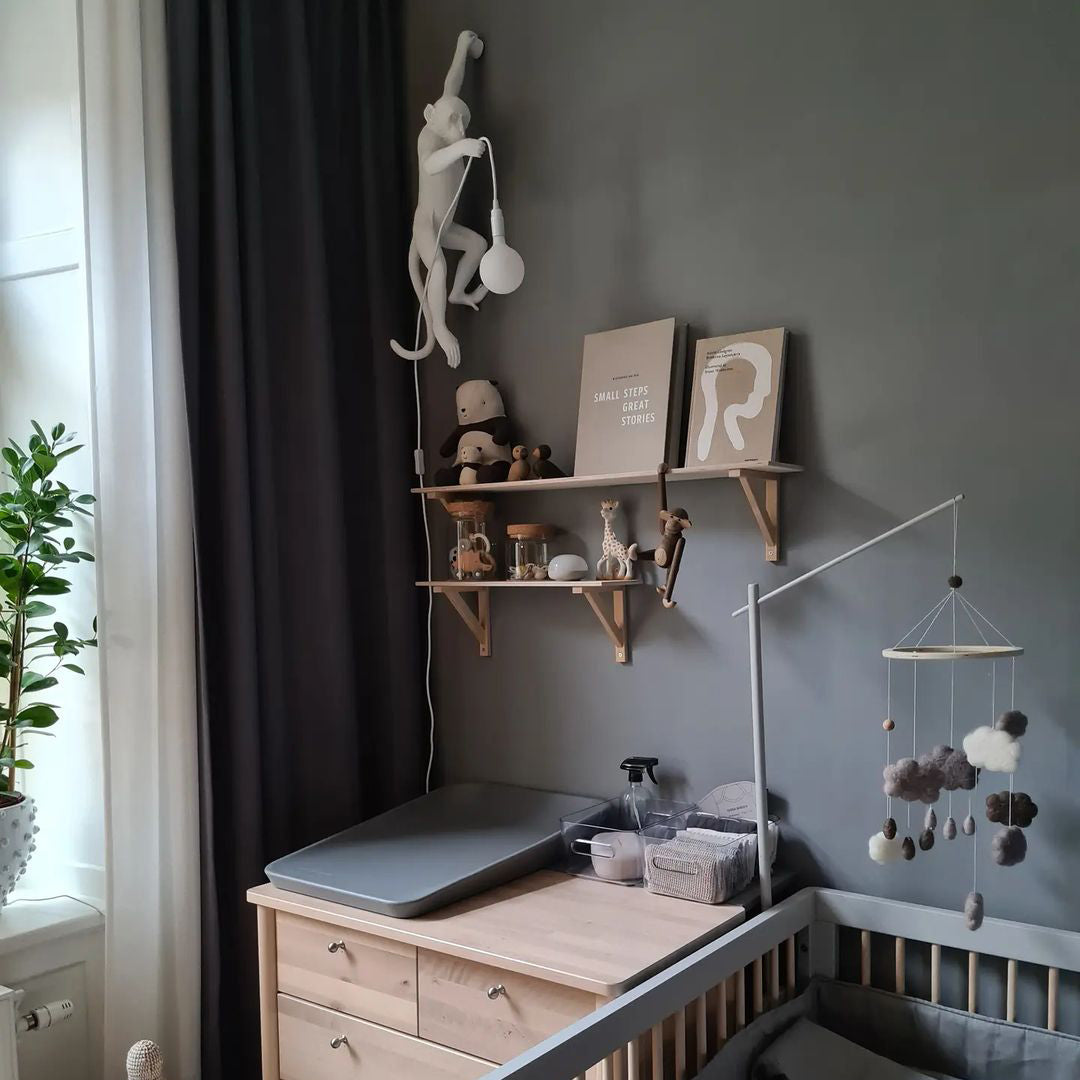 Above a changing table in a nursery with a grey wall, a white Clérachic monkey wall light hangs high with its bulb dangling.