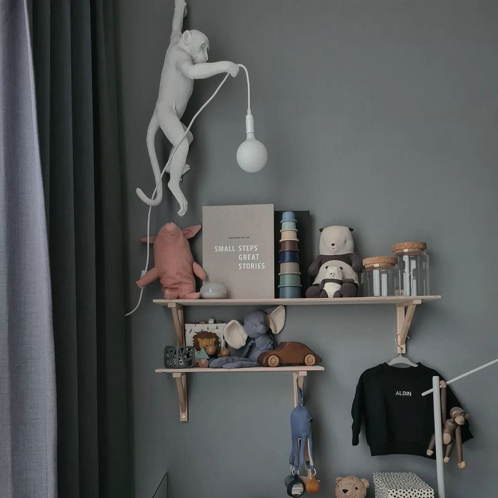 In a child's room with a dark grey wall and wooden shelves of toys, a white Clérachic monkey light hangs beside its round bulb.