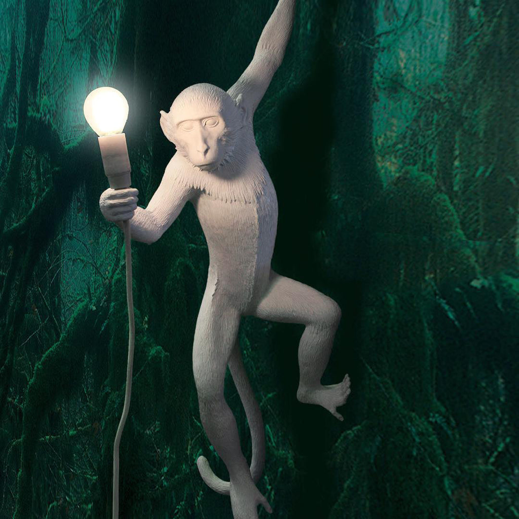 A white Clérachic monkey light holds a bright bulb against a deep green forest-print wall, one arm raised overhead.