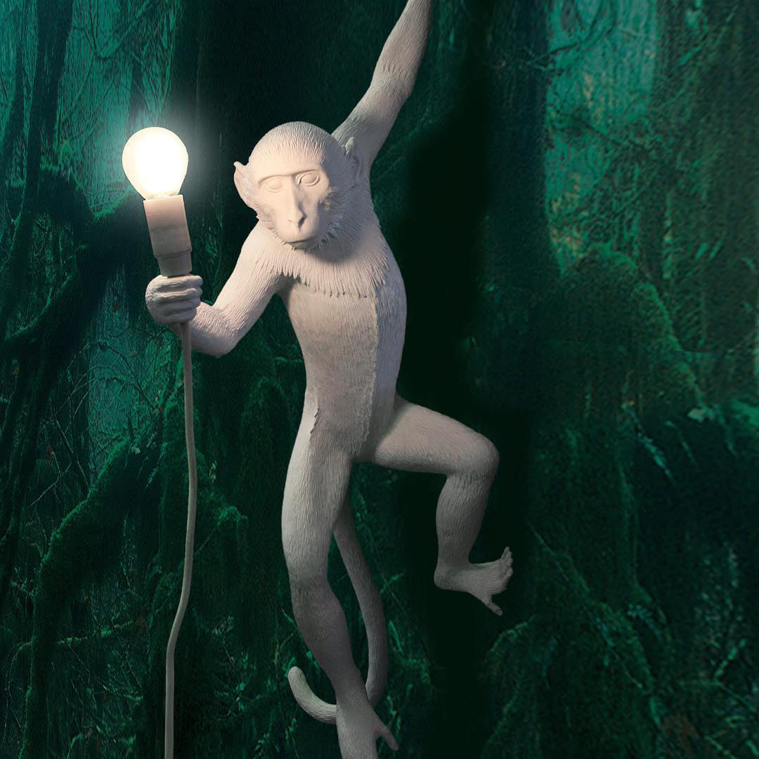 A white Clérachic monkey light holds a bright bulb against a deep green forest-print wall, one arm raised overhead.