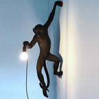 A dark bronze-toned Clérachic monkey light grips a blue-grey wall with one arm, holding a glowing bulb at its side.