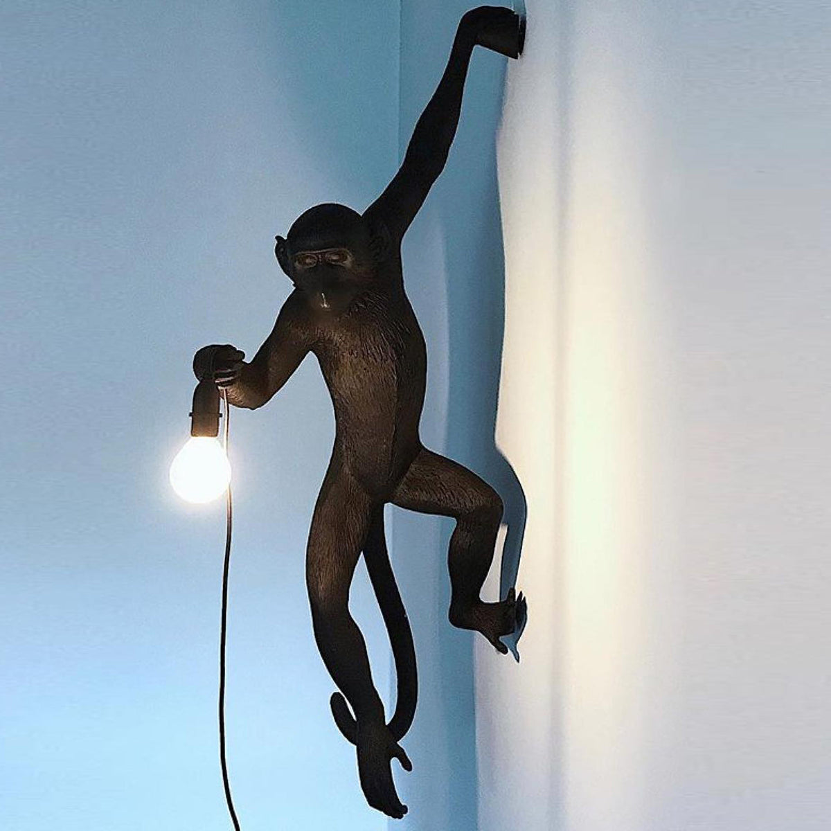 A dark bronze-toned Clérachic monkey light grips a blue-grey wall with one arm, holding a glowing bulb at its side.