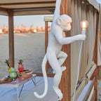 On a beach cabana, a white Clérachic monkey wall light hangs from a wooden post, a warm bulb lit in its hand.