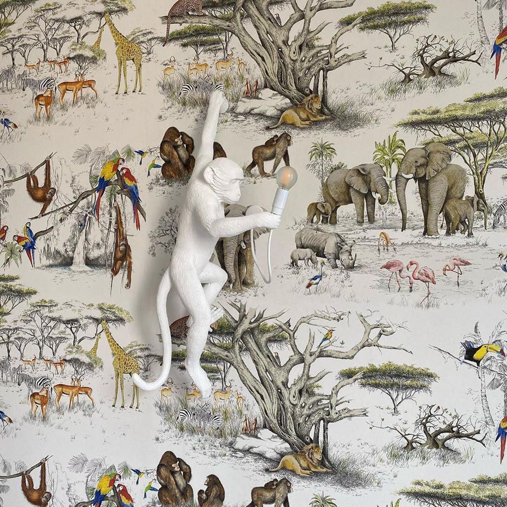 A white Clérachic monkey light stands out against a safari wallpaper of elephants, giraffes and parrots, its bulb held out.