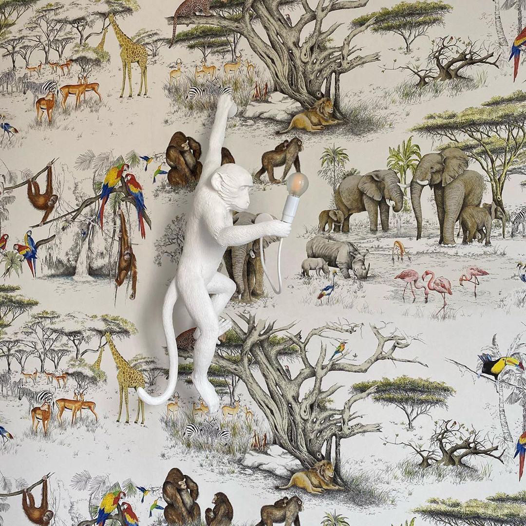A white Clérachic monkey light stands out against a safari wallpaper of elephants, giraffes and parrots, its bulb held out.