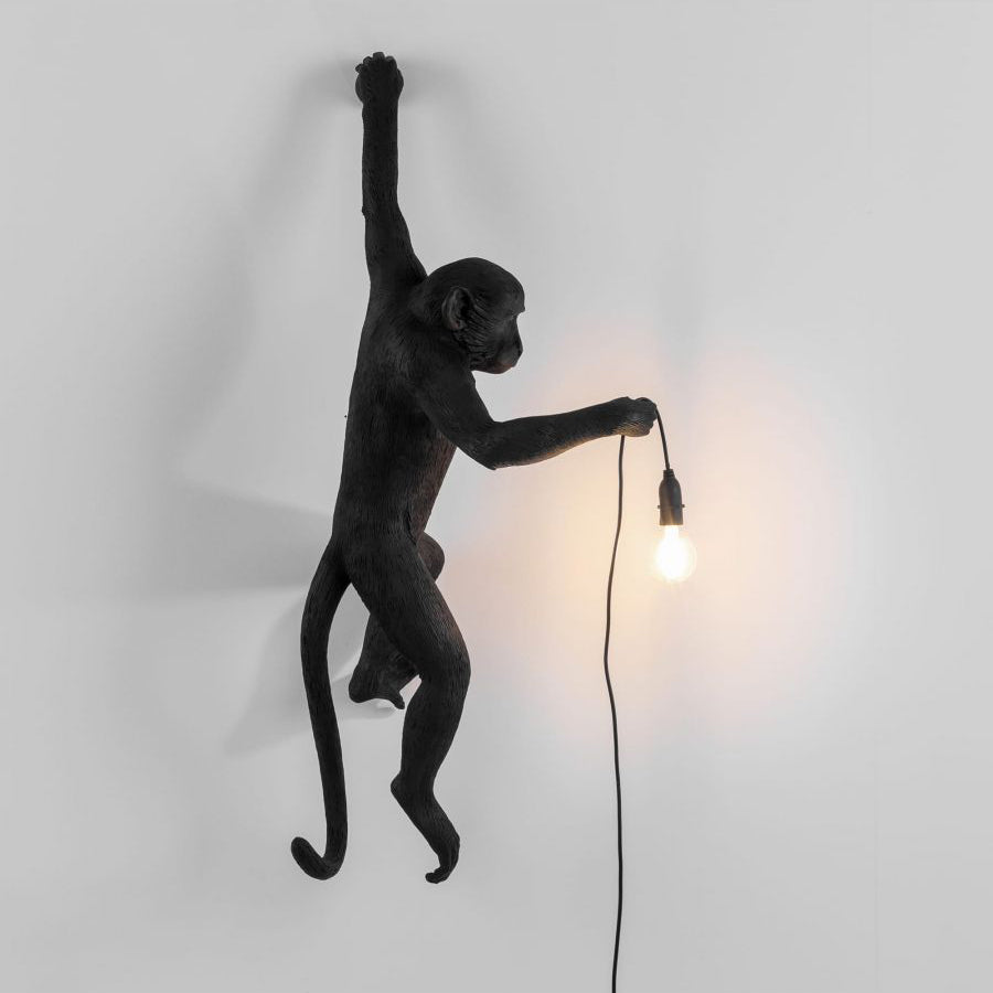 Black monkey wall light from Clérachic on a pale grey wall, one arm raised, the other holding a warm filament bulb.