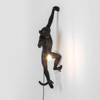 A black Clérachic monkey light hangs by one arm on a pale grey wall, its bulb glowing softly at the level of its waist.