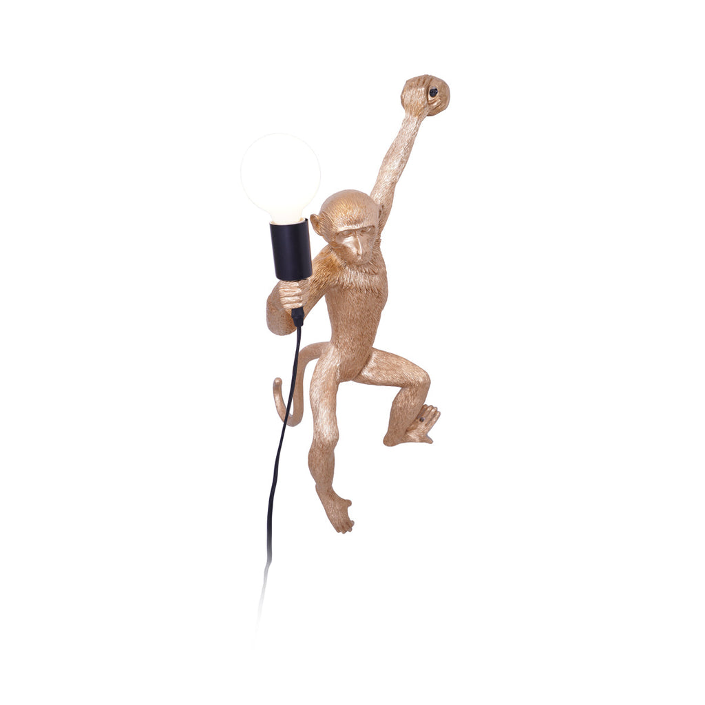 A gold Clérachic monkey wall light on a plain white backdrop, one arm gripping the top, with a black socket and white globe.