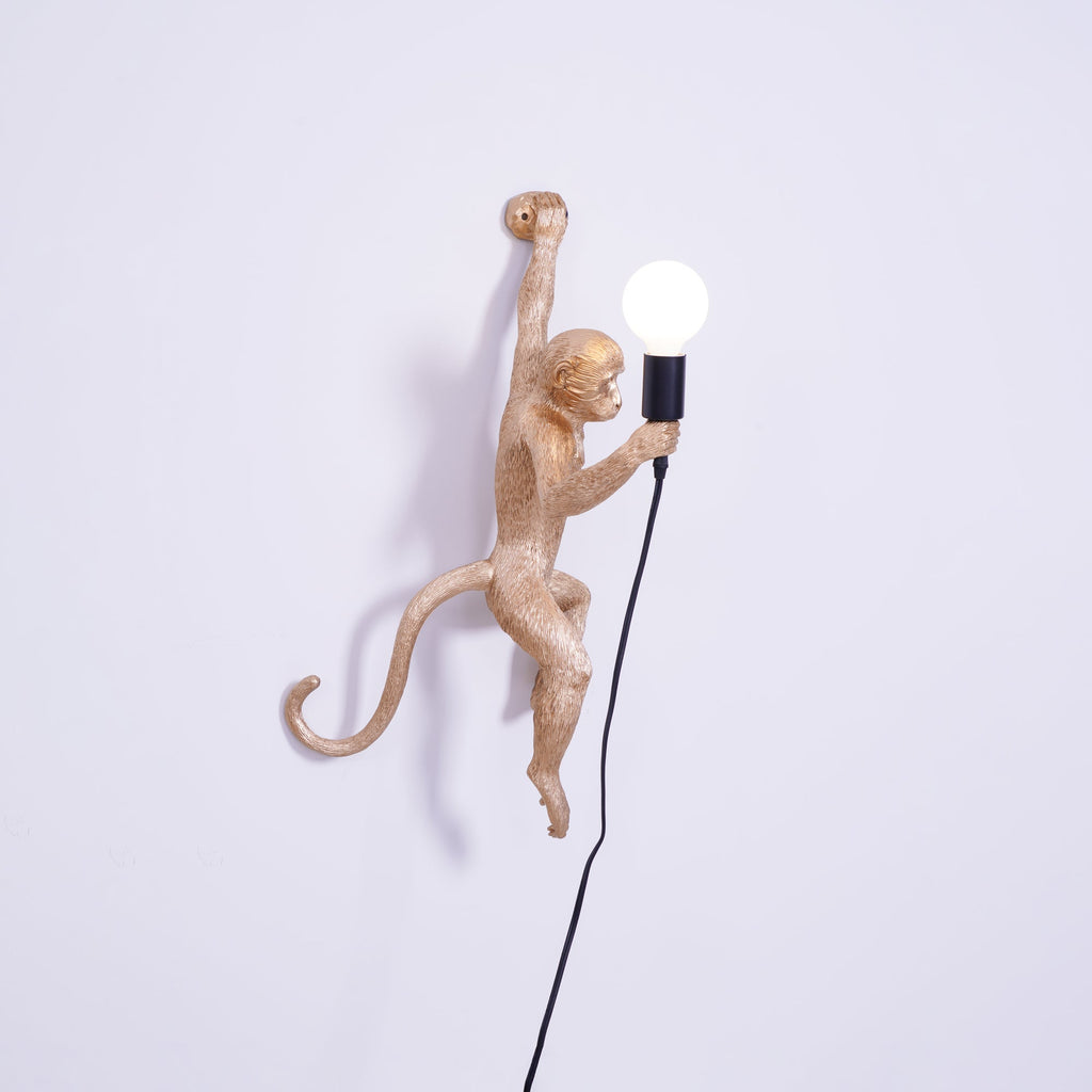 Gold monkey of this Clérachic wall light shown side-on on a lilac-white wall, its tail curled out and a black cord falling below.