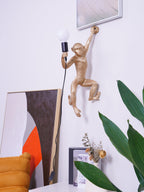 In a tidy white room, this gold Clérachic monkey light holds a globe bulb, a framed painting and a plant at its side.