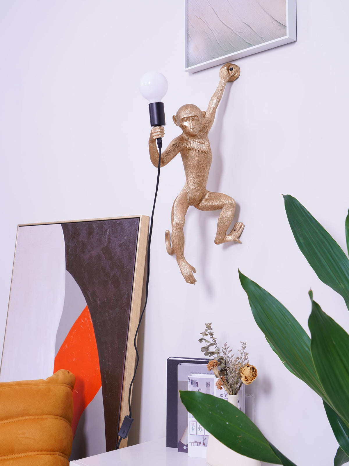 In a tidy white room, this gold Clérachic monkey light holds a globe bulb, a framed painting and a plant at its side.