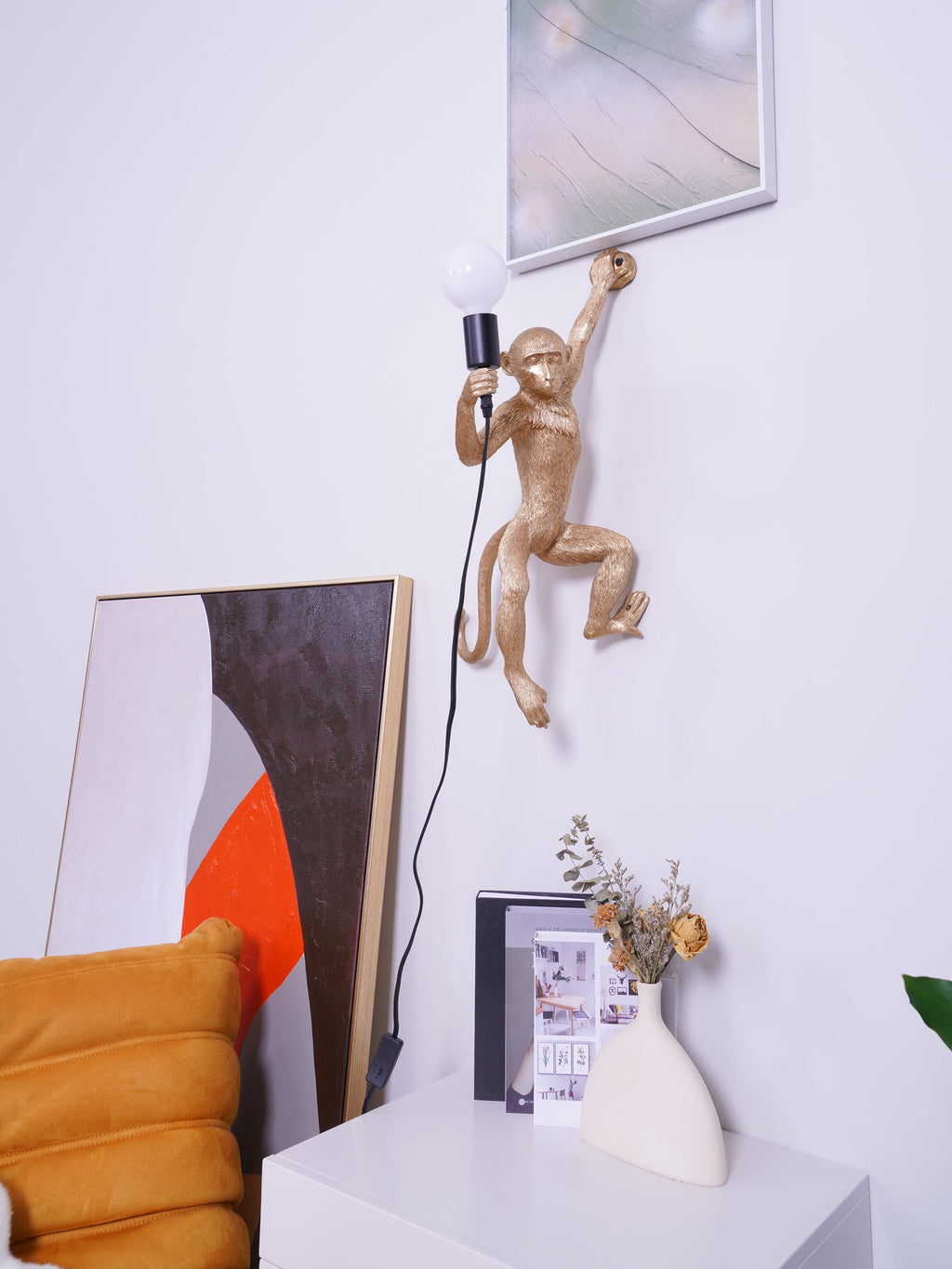 A gold Clérachic monkey wall light at the upper centre of a white wall, a framed print above it and a cabinet below.