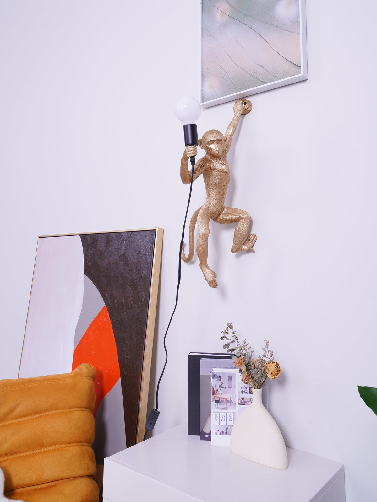 A gold Clérachic monkey wall light at the upper centre of a white wall, a framed print above it and a cabinet below.