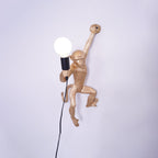 A gold Clérachic monkey wall light, close in, holding a globe bulb with its black cord hanging down the plain wall.