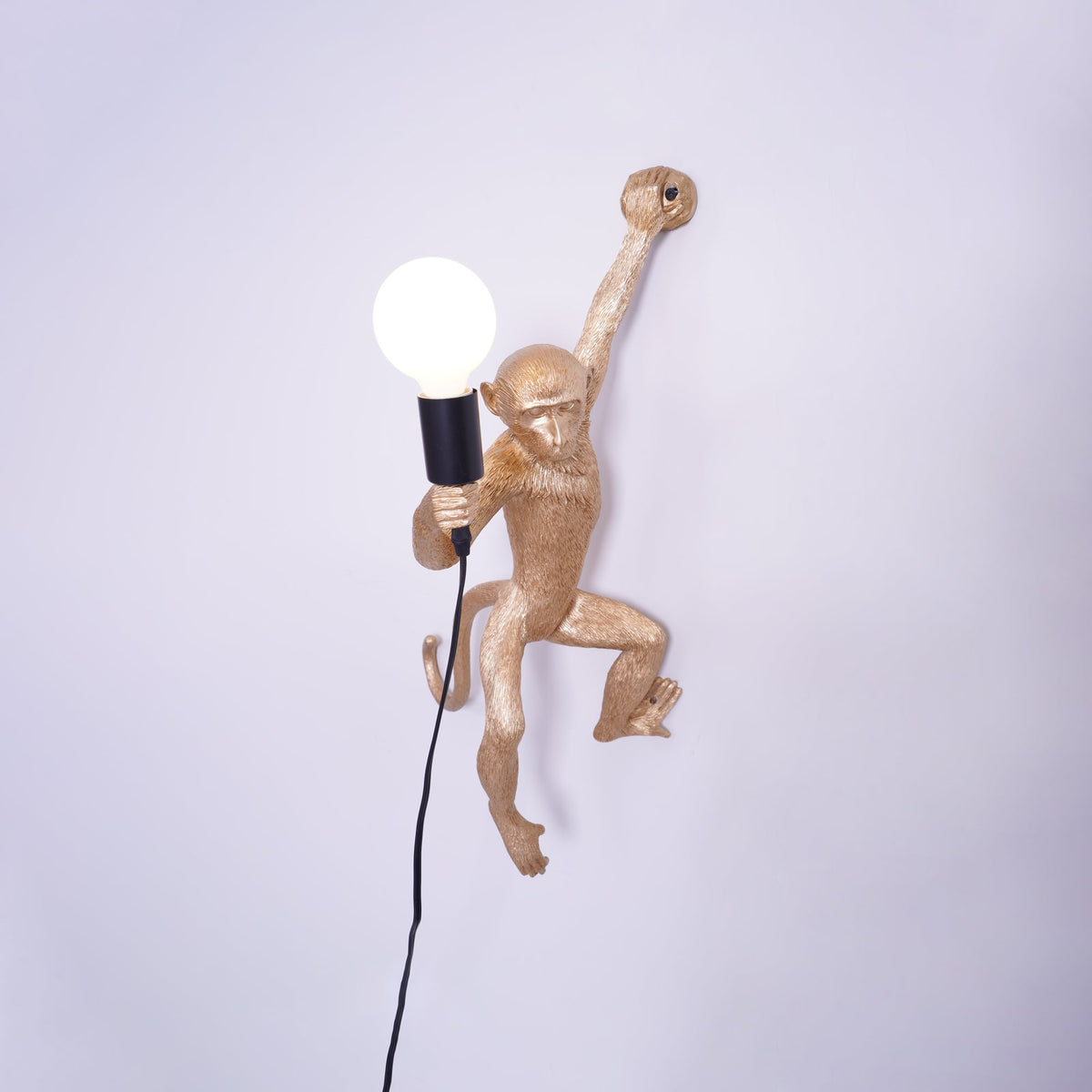 A gold Clérachic monkey wall light, close in, holding a globe bulb with its black cord hanging down the plain wall.