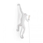 A white Clérachic monkey light almost vanishes into a white backdrop, one arm up, with a small bulb held out to the right.