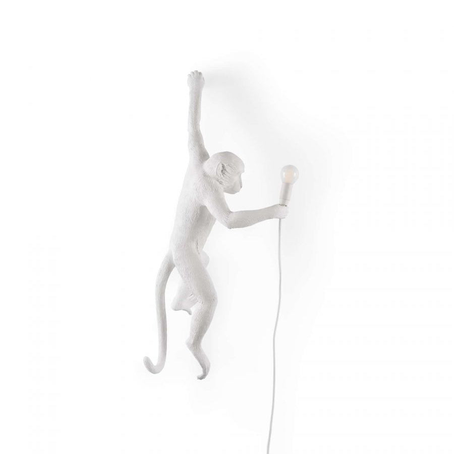 White resin monkey wall light from Clérachic on a white backdrop, leaning forward with a small bulb in its outstretched hand.