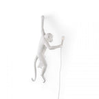 White resin monkey wall light from Clérachic on a white backdrop, leaning forward with a small bulb in its outstretched hand.