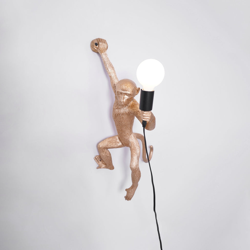 A gold Clérachic monkey light close up on a pale grey wall, its large globe bulb lit and a black cord falling behind.