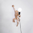 A gold Clérachic monkey light close up on a pale grey wall, its large globe bulb lit and a black cord falling behind.