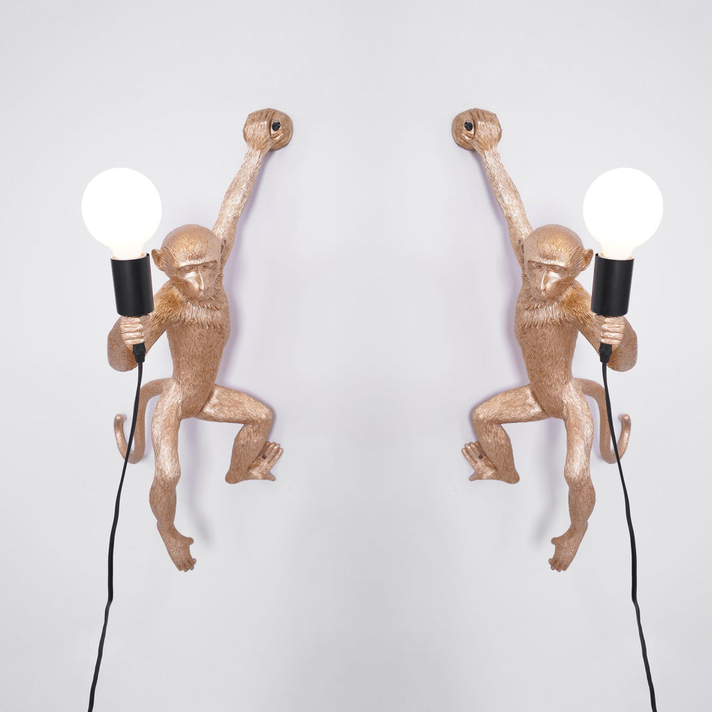 A pair of gold Clérachic monkey wall lights, left and right mirrored, each holding a white globe bulb on a pale grey wall.
