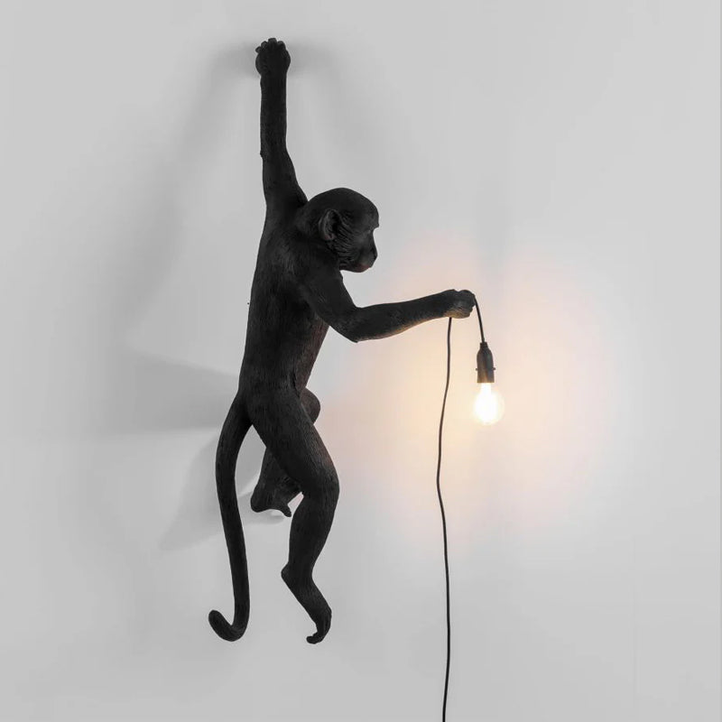 A black Clérachic monkey light with its arm raised high and a warm filament bulb glowing at the end of its cord to the right.